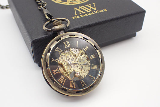 Antique bronze mechanical pocket watch with an open-face design and a gear-textured bezel. The dial features a black outer ring with gold Roman numerals and a large central cut-out revealing an intricate golden hand-wind mechanical movement. Resting beside a black MW gift box.