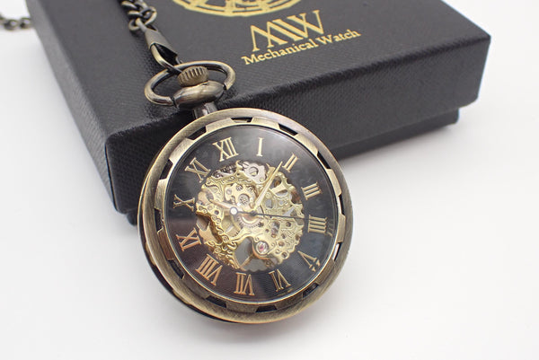 Antique bronze mechanical pocket watch with an open-face design and a gear-textured bezel. The dial features a black outer ring with gold Roman numerals and a large central cut-out revealing an intricate golden hand-wind mechanical movement. Resting beside a black MW gift box.