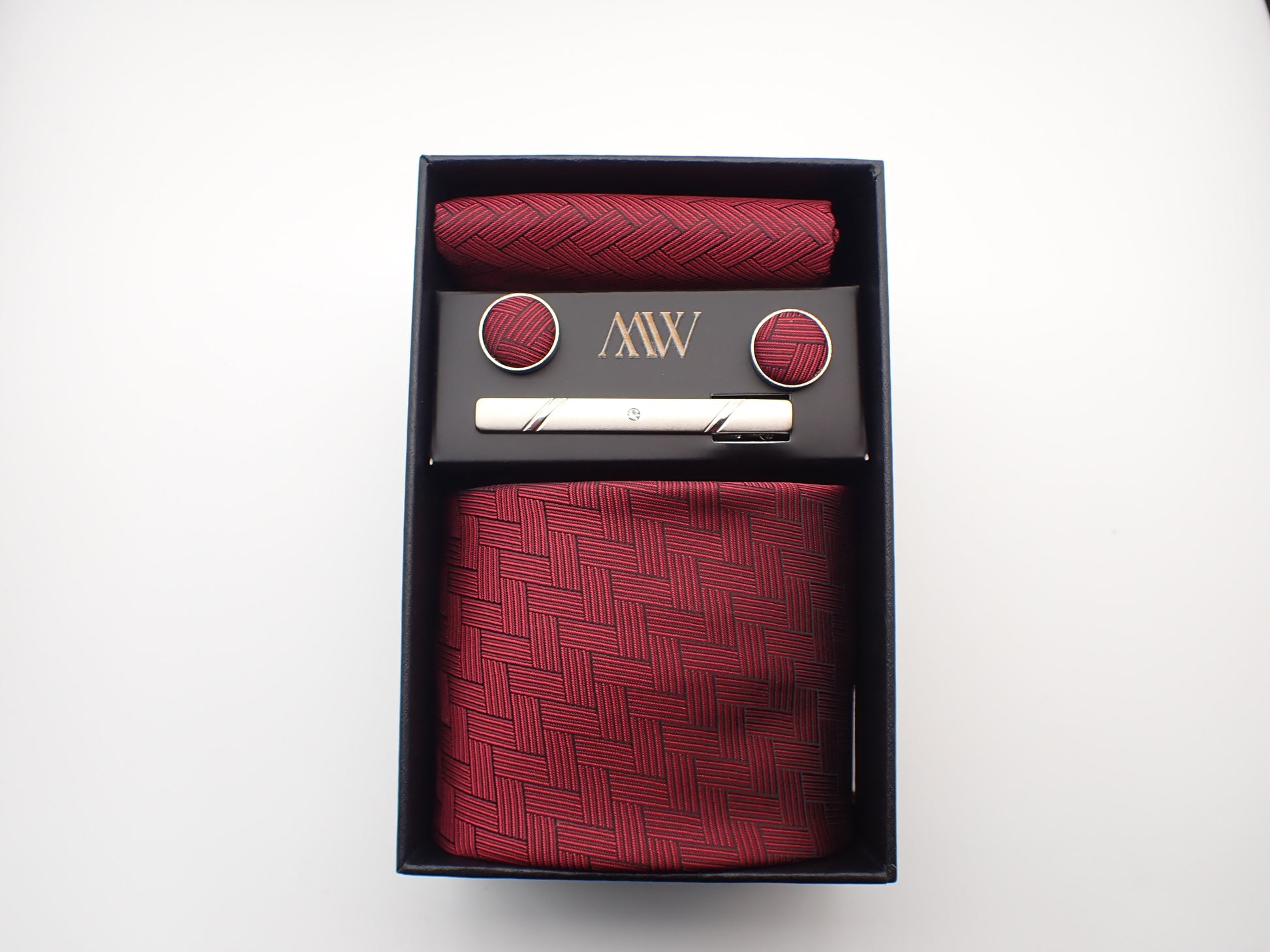 A black gift box containing a dark burgundy red men's tie with a geometric basketweave woven texture, matching pocket square, cufflinks, and a tie clip.