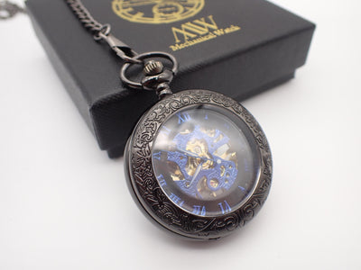 Gunmetal black mechanical pocket watch featuring an intricately engraved floral rim and an open face that reveals a vibrant blue skeleton movement and blue Roman numerals, resting on a black MW premium gift box.