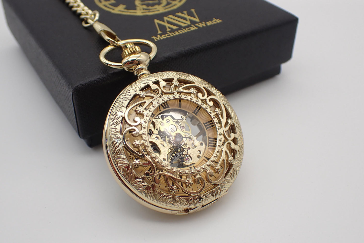 A luxurious yellow gold mechanical pocket watch featuring a highly ornate floral filigree cut-out cover and engraved rim. The dial reveals black Roman numerals on a gold background and an exposed golden hand-wind mechanical movement. Resting beside a black MW premium gift box.