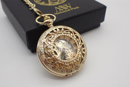 A luxurious yellow gold mechanical pocket watch featuring a highly ornate floral filigree cut-out cover and engraved rim. The dial reveals black Roman numerals on a gold background and an exposed golden hand-wind mechanical movement. Resting beside a black MW premium gift box.