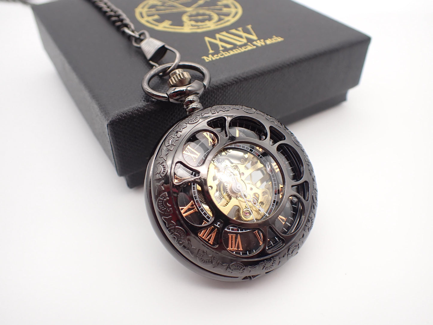 Gunmetal black skeleton pocket watch with a cut-out front cover, revealing rose gold Roman numerals and a golden hand-wind mechanical movement. Resting next to a black MW premium gift box.