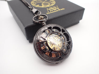Gunmetal black skeleton pocket watch with a cut-out front cover, revealing rose gold Roman numerals and a golden hand-wind mechanical movement. Resting next to a black MW premium gift box.