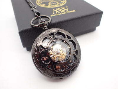 Dark gunmetal mechanical pocket watch with a circular cut-out cover. The skeleton dial reveals rose gold Roman numerals, an inner 24-hour track, elegant white ornate hands, and a visible gold-toned movement. Resting beside a black MW gift box.