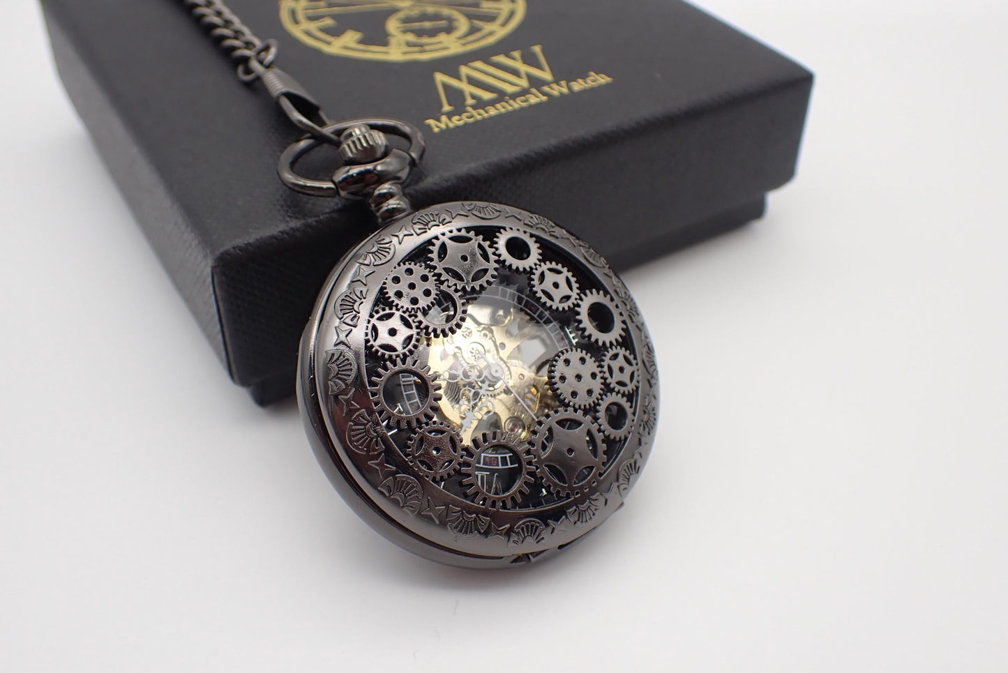 Dark gunmetal mechanical pocket watch featuring a unique steampunk multi-gear cut-out front cover and engraved rim. The skeleton design reveals a central golden hand-wind mechanical movement. Resting next to a black MW premium gift box.