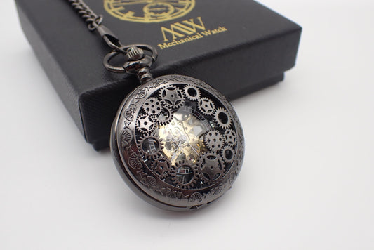Dark gunmetal mechanical pocket watch featuring a unique steampunk multi-gear cut-out front cover and engraved rim. The skeleton design reveals a central golden hand-wind mechanical movement. Resting next to a black MW premium gift box.