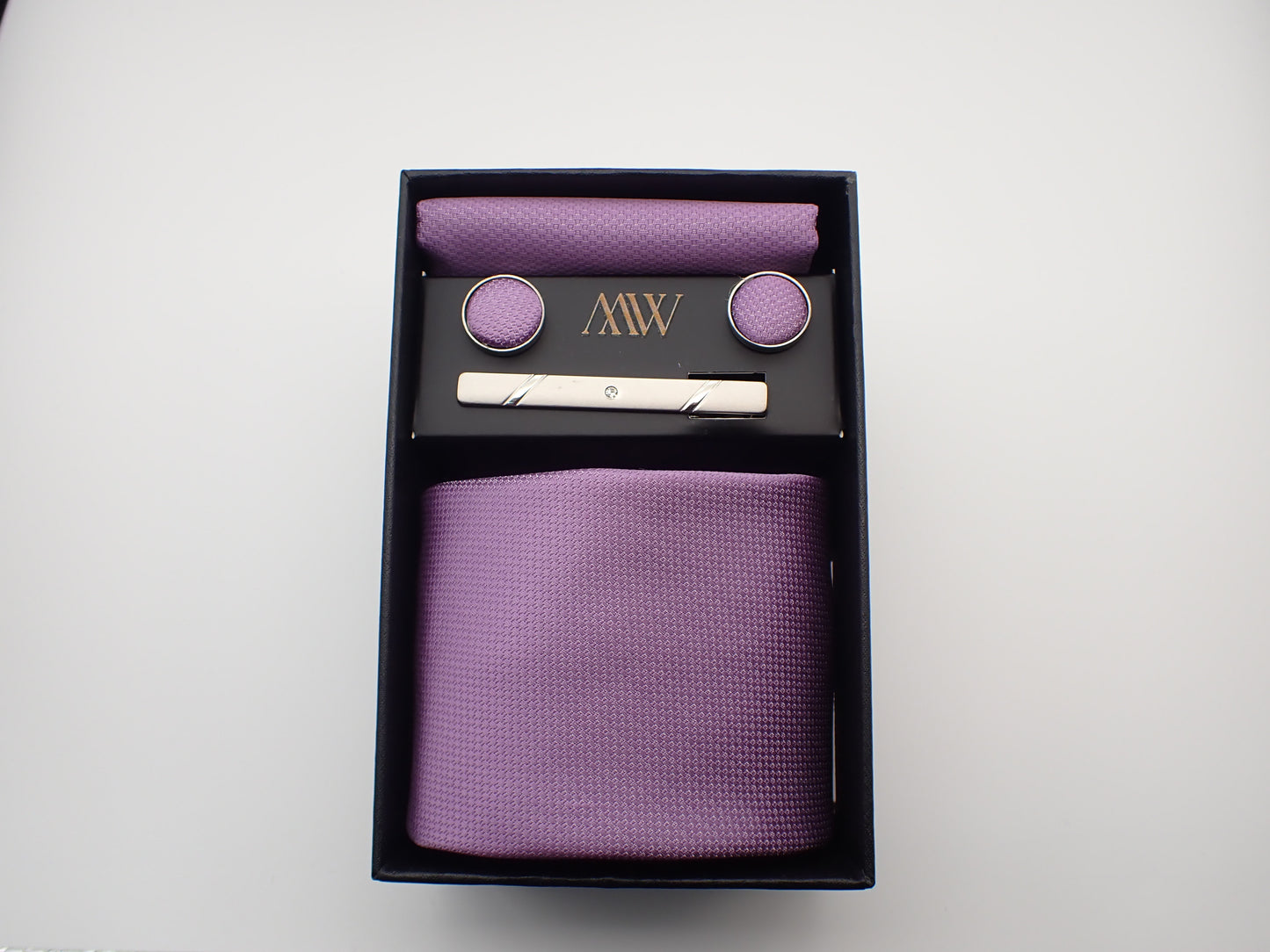 A black gift box displaying a light lavender purple textured men's necktie, matching pocket square, round cufflinks, and a silver tie clip.