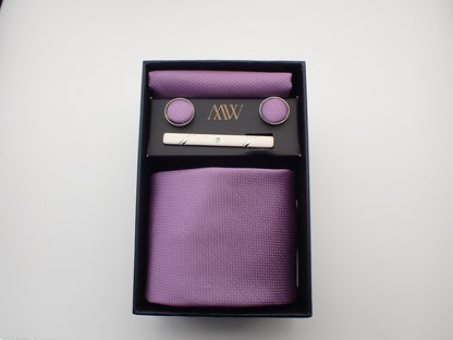 A black gift box displaying a light lavender purple textured men's necktie, matching pocket square, round cufflinks, and a silver tie clip.