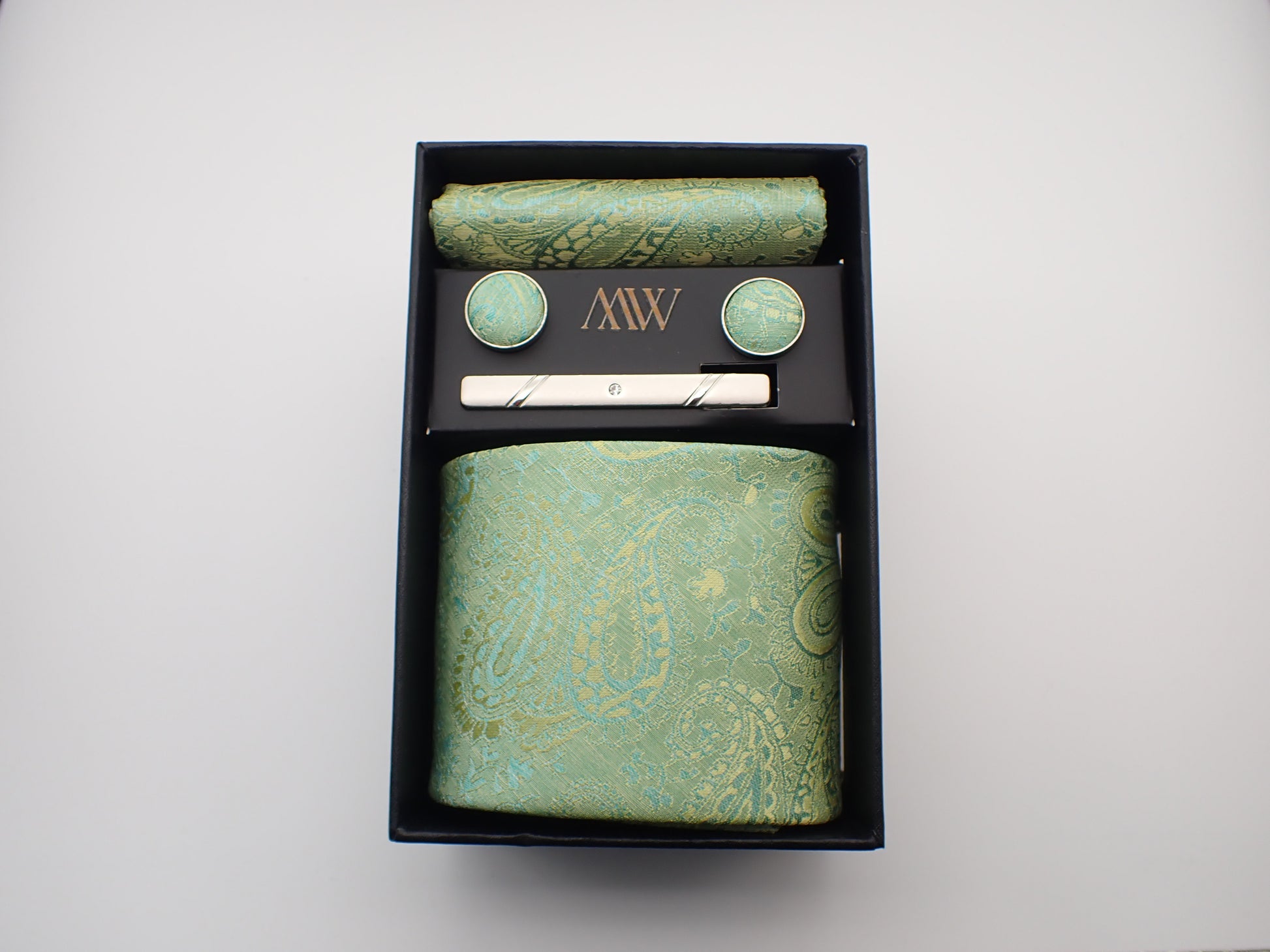 A black gift box presenting a light green and gold paisley floral jacquard patterned men's necktie, matching pocket square, cufflinks, and a tie clip.