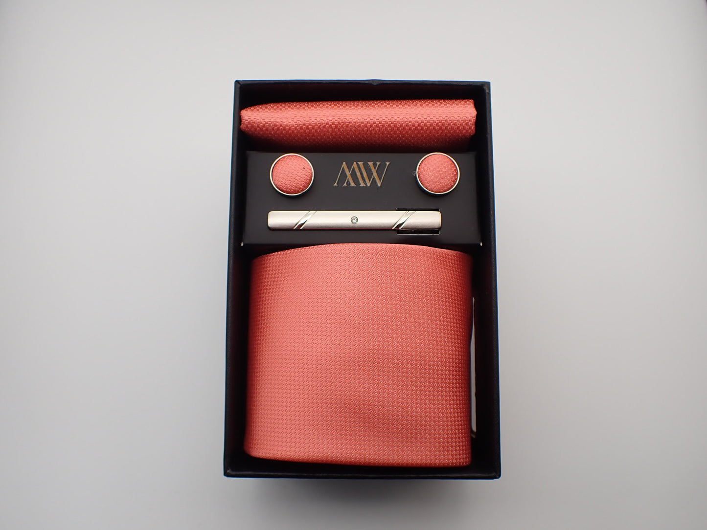 A black gift box containing a coral pink micro-woven textured men's necktie, matching pocket square, round cufflinks, and a silver tie clip.