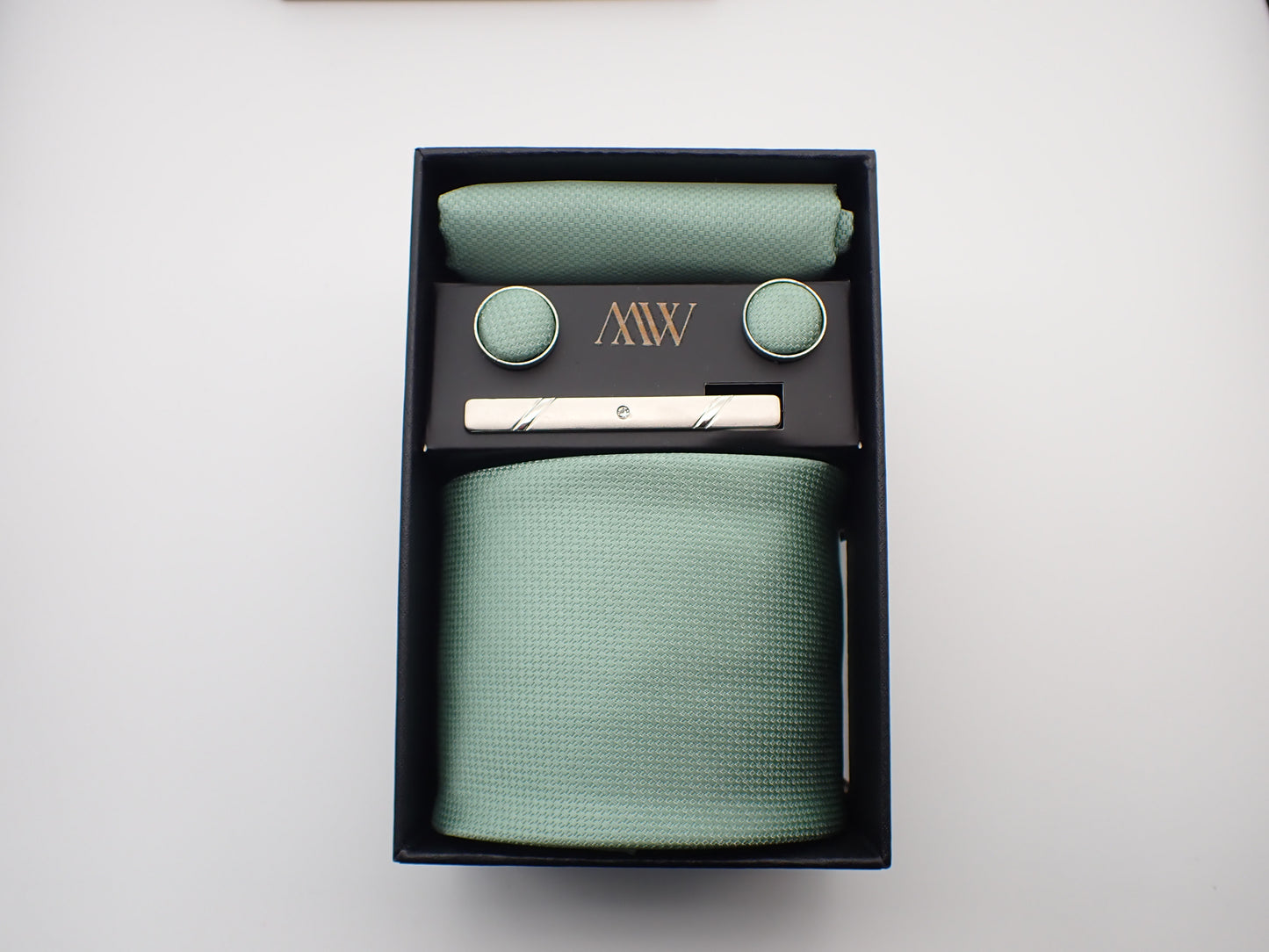 A luxury black gift box containing a pastel mint green micro-woven men's necktie, matching pocket square, cufflinks, and a silver tie clip.