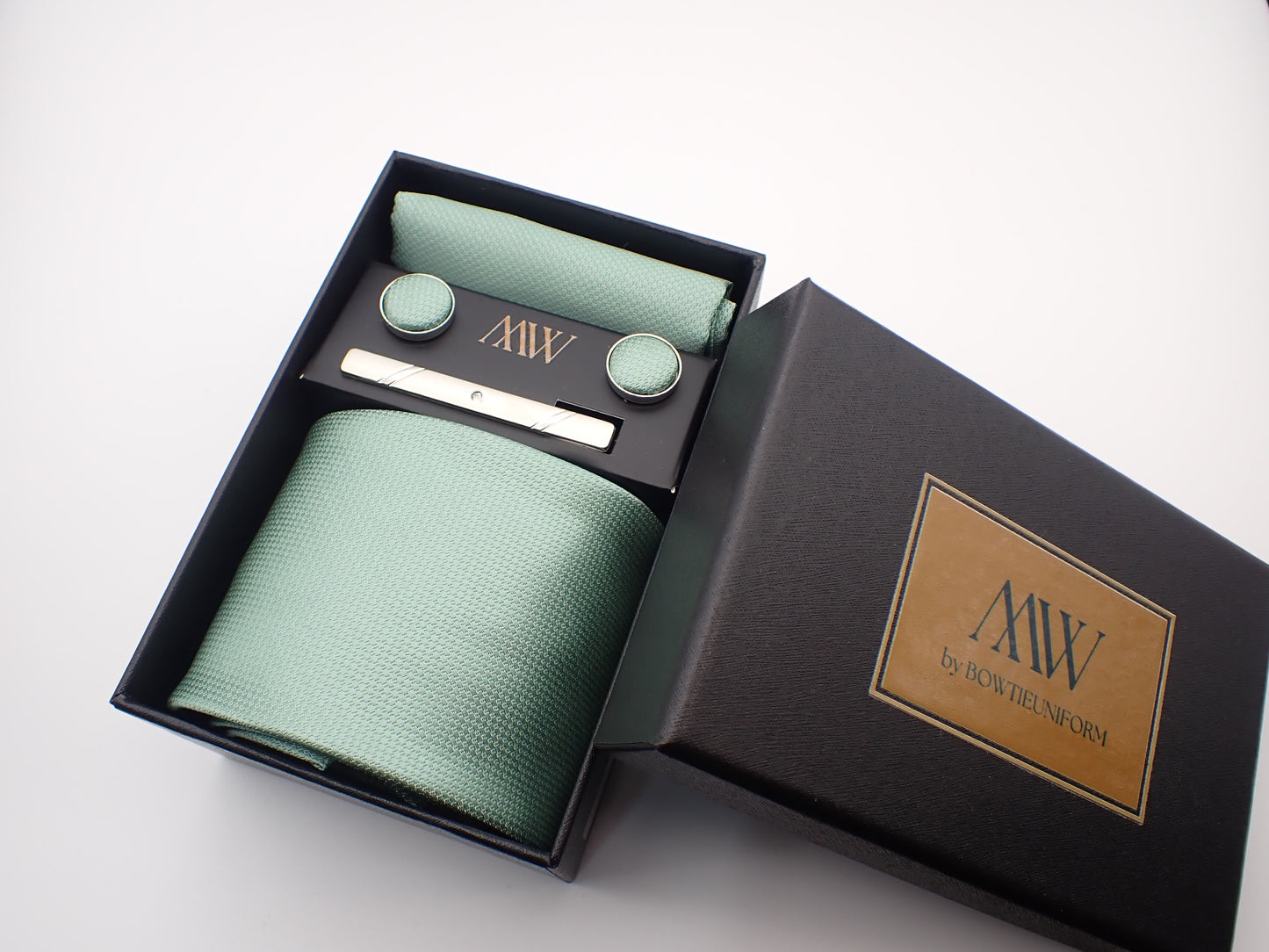 A luxury black gift box containing a pastel mint green micro-woven men's necktie, matching pocket square, cufflinks, and a silver tie clip.