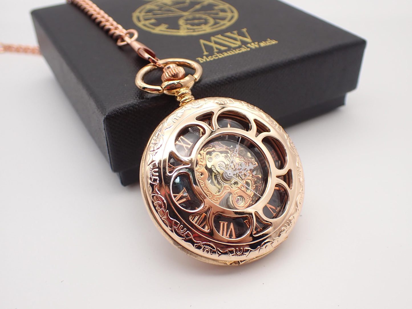 An elegant rose gold mechanical pocket watch with complex circular and line-pattern engravings on the rim. The front cover is intricately cut out, revealing a black dial with rose gold Roman numerals and a central gold and rose gold-toned mechanical movement. Rest beside a closed black MW premium gift box with its gold logo on a clean white surface.
