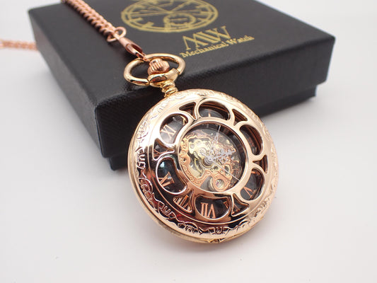 An elegant rose gold mechanical pocket watch with complex circular and line-pattern engravings on the rim. The front cover is intricately cut out, revealing a black dial with rose gold Roman numerals and a central gold and rose gold-toned mechanical movement. Rest beside a closed black MW premium gift box with its gold logo on a clean white surface.