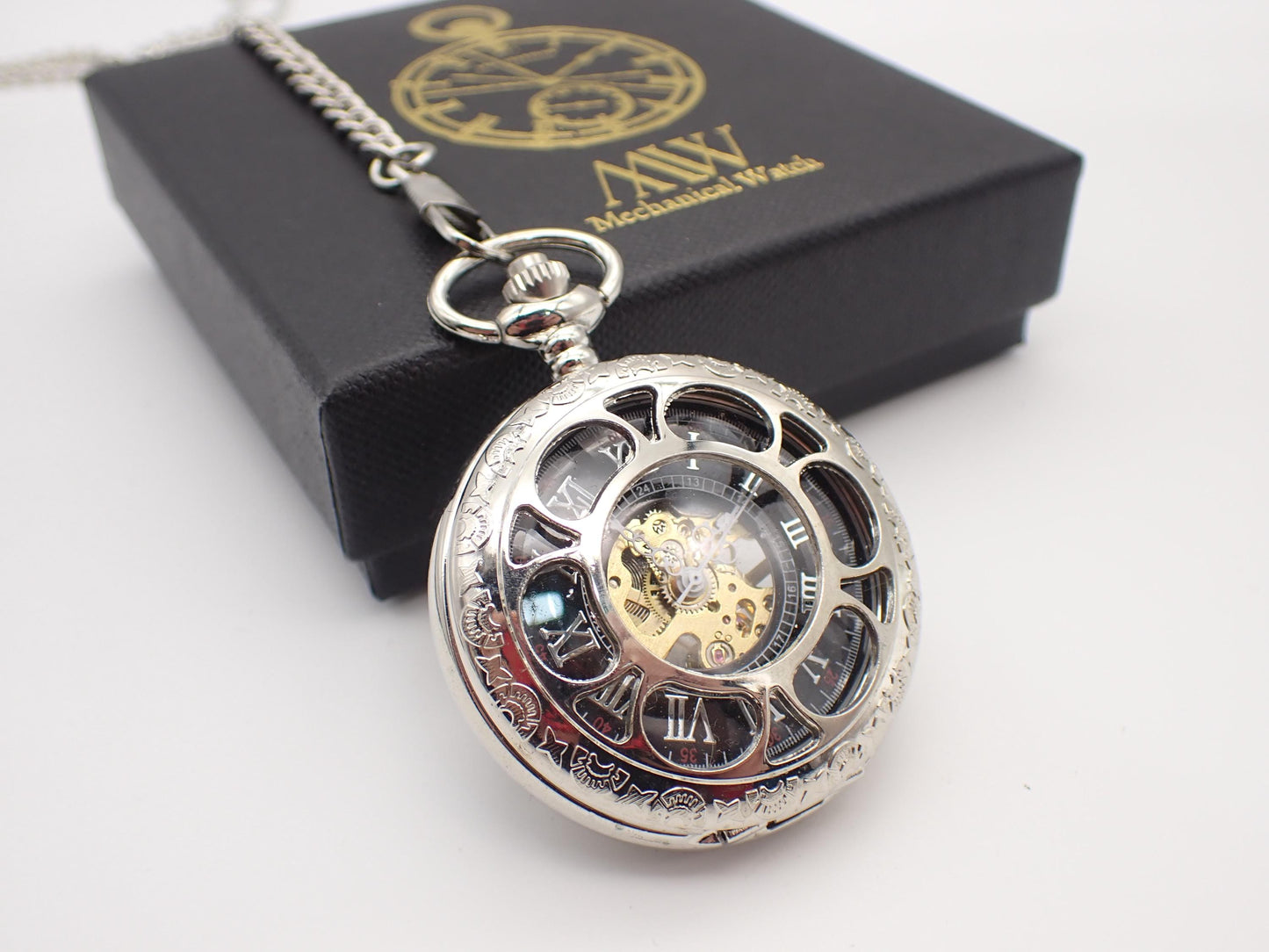 Polished silver mechanical pocket watch with intricate floral engravings on the rim and a circular cut-out cover, revealing a black dial with silver Roman numerals and a central gold and silver-toned mechanical movement. Resting beside a black MW gift box.