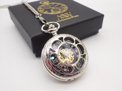 Polished silver mechanical pocket watch with intricate floral engravings on the rim and a circular cut-out cover, revealing a black dial with silver Roman numerals and a central gold and silver-toned mechanical movement. Resting beside a black MW gift box.