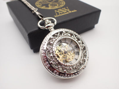 Silver mechanical pocket watch featuring an ornate filigree floral cutout cover revealing a gold-tone skeleton movement, placed on a black MW premium gift box.