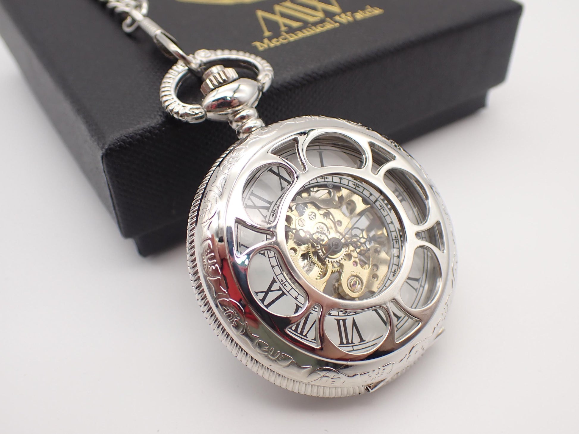 Silver mechanical pocket watch with an open-face skeleton dial revealing gold gears and black Roman numerals, resting on a black premium MW gift box.