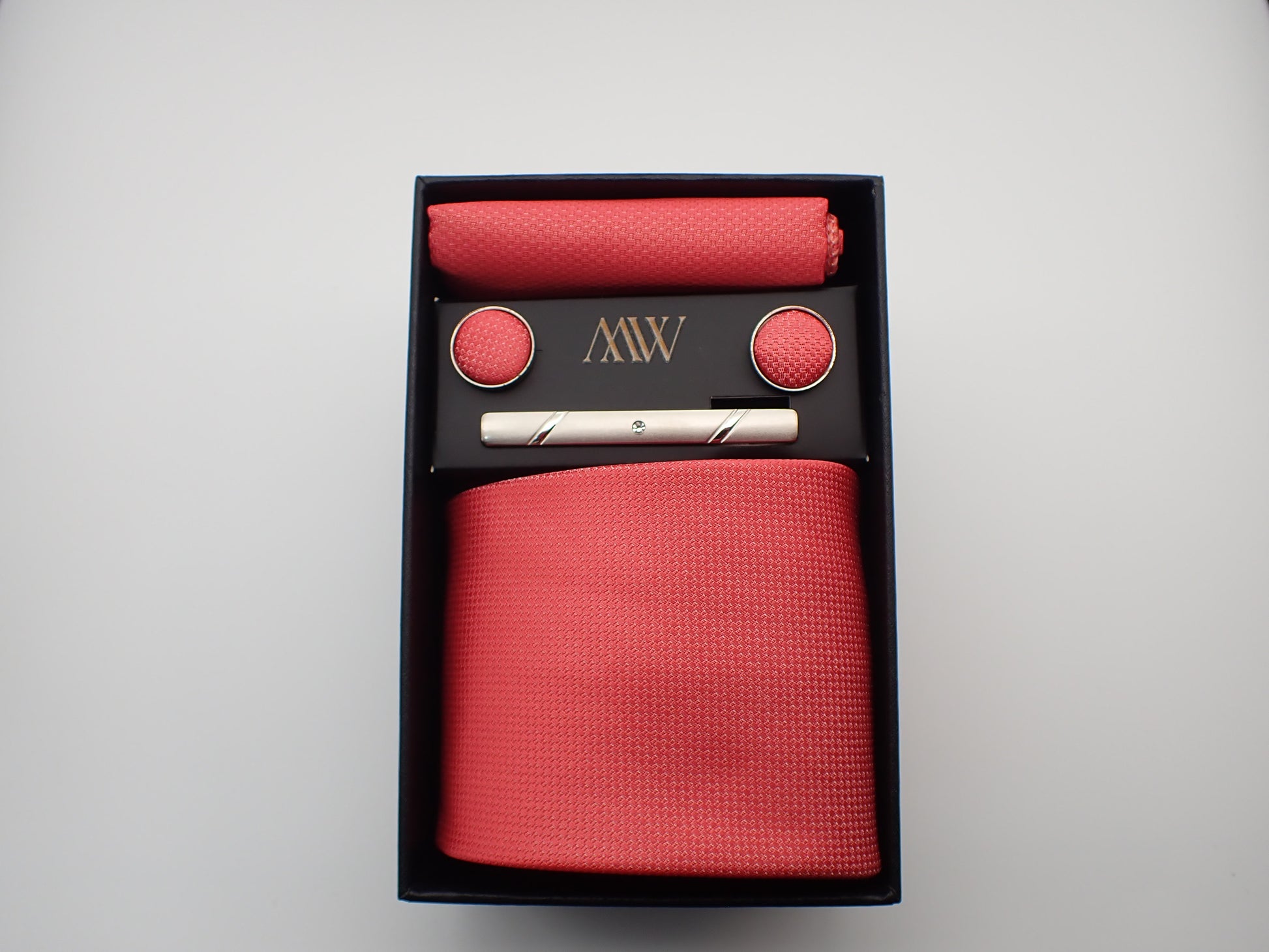A black gift box featuring a bright watermelon pink textured men's necktie, a matching pocket square, round cufflinks, and a silver tie bar.