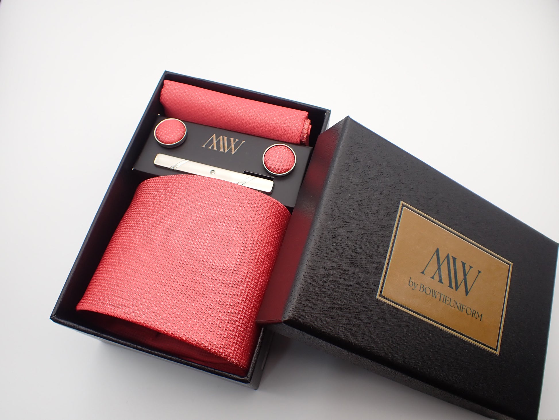 A black gift box featuring a bright watermelon pink textured men's necktie, a matching pocket square, round cufflinks, and a silver tie bar.